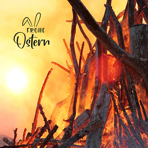 Ein großes, loderndes Feuer brennt in der Mitte des Bildes, umgeben von Holzstämmen. Im oberen Teil steht in geschwungener Schrift „Frohe Ostern“. Die Szene vermittelt eine festliche Atmosphäre und symbolisiert das Osterfeuer.