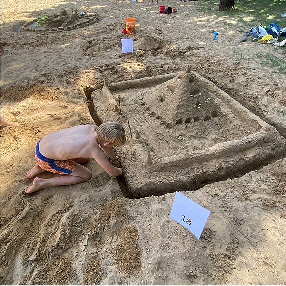 Ein Kind kniet im Sand und baut eine aufwendige Sandburg: quadratische Anlage mit Mauern, Zinnen und umlaufendem Graben, in der Mitte eine hohe pyramidenförmige Spitze. Der Fokus liegt auf dem Bauprozess und der detailreichen Burg am Strand.