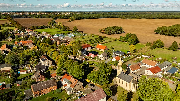 Eine Luftaufnahme eines kleinen Dorfes umgeben von Feldern und Wäldern. Im Vordergrund sind zahlreiche Häuser mit Dächern in verschiedenen Farben sowie eine Kirche sichtbar. Im Hintergrund erstreckt sich eine grüne Landschaft mit Bäumen und weitläufigen Feldern.