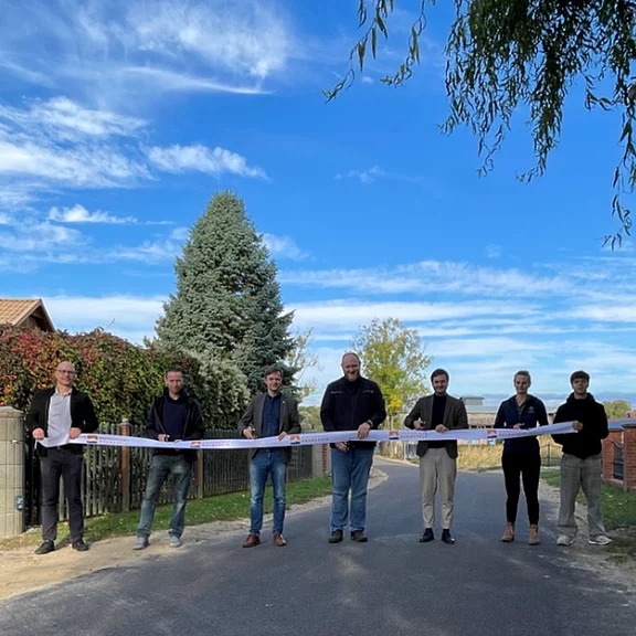 Acht Personen stehen quer über einer Dorfstraße und halten ein langes weißes Band; in der Mitte wird es mit Scheren durchtrennt. Zeremonielle Eröffnung im Freien bei blauem Himmel, ländliche Umgebung mit Bäumen und Häusern im Hintergrund.