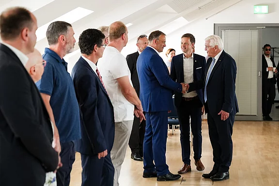 Eine Gruppe von Personen steht in einem Raum, während zwei Männer sich die Hände schütteln. Die Atmosphäre wirkt förmlich und freundlich, der Raum ist hell erleuchtet und modern eingerichtet. Im Hintergrund sind einige Teilnehmer, die den Moment beobachten.