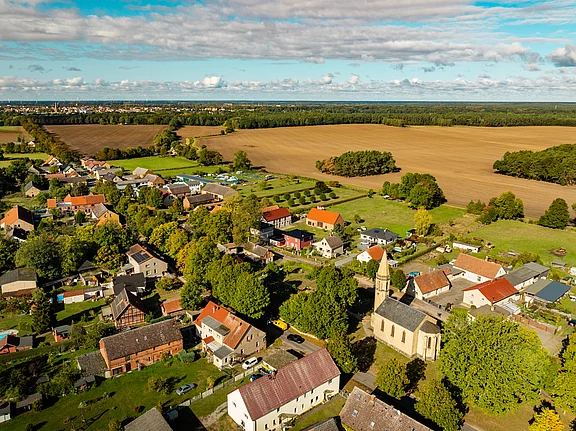 Luftaufnahme eines kleinen Dorfes mit mehreren Häusern, einer Kirche und umgebenden Feldern. Im Hintergrund sind Bäume und ein Weizenfeld zu sehen. Der Himmel ist teilweise bewölkt, mit einer klaren blauen Fläche. Die ländliche Umgebung vermittelt eine friedliche Atmosphäre.