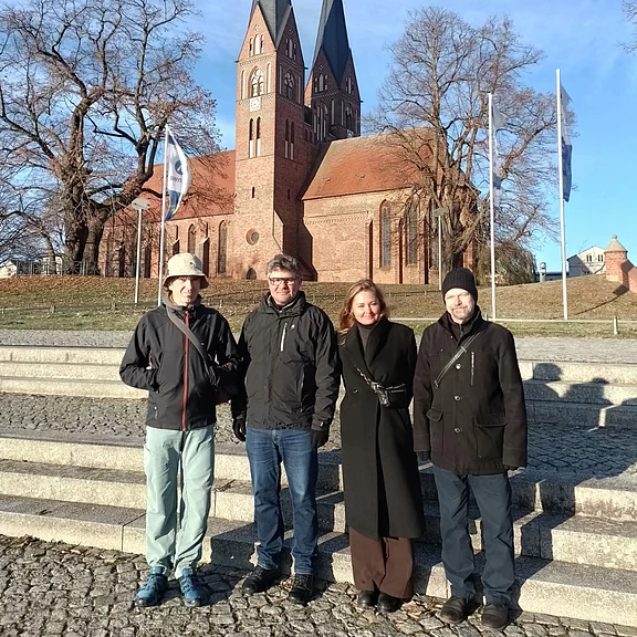 Vier Personen stehen auf steinernen Stufen und schauen in die Kamera. Sie tragen Winterkleidung. Hinter ihnen erhebt sich eine große Backsteinkirche mit zwei schlanken Türmen. Klarer Himmel, karge Bäume.