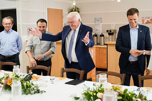 Vier Männer stehen an einem Tisch, der mit Blumen und Getränken dekoriert ist. Der Mann in der Mitte, mit Brille und Anzug, gestikuliert einladend. Die anderen Männer schauen aufmerksam, während sie sich um den Tisch gruppieren. Die Stimmung wirkt freundlich und gesellig.