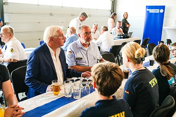 Eine Gruppe von Menschen sitzt an einem Tisch in einem hellen Raum. Zwei Männer in Anzügen führen ein Gespräch. Auf dem Tisch stehen Gläser mit Getränken. Im Hintergrund sind weitere Personen und eine große Banner mit dem Logo einer Organisation zu sehen.