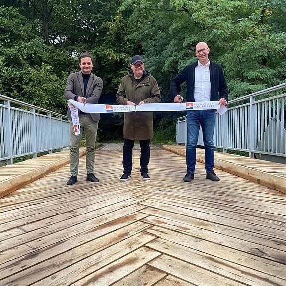 Drei Männer stehen auf einer neuen Fußgängerbrücke und halten ein Einweihungsband mit Logos gespannt. Holzbohlen und Metallgeländer rahmen die Szene. Feierliche Eröffnung der Brücke im Freien.