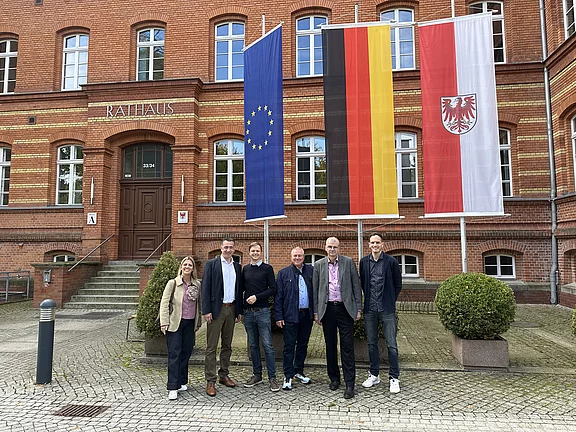 Sechs Personen stehen vor einem historischen Rathaus aus Backstein. Daneben wehen drei große Fahnen: Europäische Union, Deutschland und Brandenburg. Der Eingang mit der Aufschrift Rathaus ist sichtbar. Die Szene wirkt wie ein offizieller Besuch oder Fototermin.
