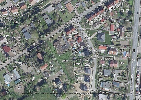 Luftbild eines Wohngebiets mit eingezeichneten Grundstücksgrenzen und Flurstücksnummern. Mehrere Einfamilienhäuser, Gärten und Straßen sind erkennbar; die Parzellen sind weiß umrandet und nummeriert.