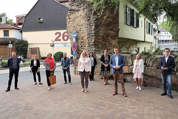 Gruppe von zehn Erwachsenen posiert im Freien auf gepflasterter Fläche. Sie stehen mit Abstand zueinander, überwiegend in Business‑Kleidung, und blicken zur Kamera. Im Hintergrund eine alte Steinmauer und Häuser. Szene wirkt wie ein offizielles Treffen oder Fototermin.