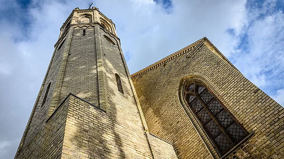 Ein historischer Gebäudeturm in hellem Ziegelstein, hoch aufragend unter einem bewölkten Himmel. Die Architektur zeigt eine Mischung aus gotischen und modernen Elementen, mit einem markanten Fenster auf der Seitenfassade. Schatten spielen auf der Oberfläche, was der Szene eine dramatische Wirkung verleiht.
