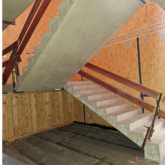 Rohbau mit Betontreppe im Inneren: zwei gegenläufige Treppenläufe, provisorisch mit Holzgeländern und Stützen gesichert. OSB‑Wände, unfertiger Boden. Bild zeigt Baustellensituation des Treppenhauses.