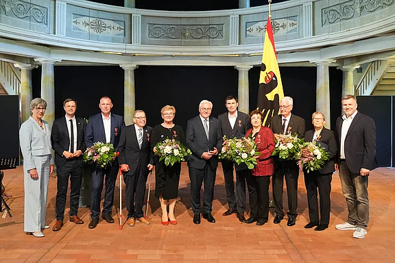 Eine Gruppe von neun Personen steht in einem festlichen Raum. Sie halten alle Blumensträuße und tragen verschiedene formelle Kleidung. Vorne befindet sich ein Mann mit einem Stock, neben ihm eine Frau mit einer roten Blume an ihrer Kleidung. Im Hintergrund ist eine Flagge zu sehen.
