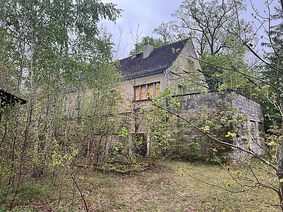 Verwildertes, zweistöckiges Haus am Waldrand: gealterte Fassade, dunkles Ziegeldach. Dichte Büsche und junge Bäume überwuchern das Grundstück und verdecken Teile des Gebäudes. Gesamtwirkung: verlassen und verfallen.