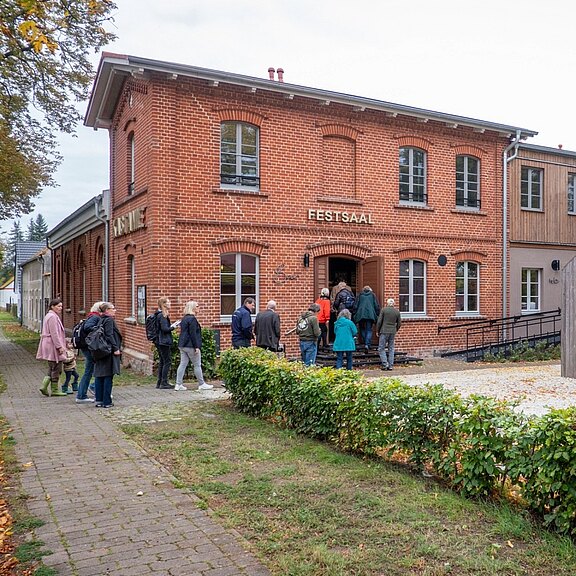 Mehrere Menschen versammeln sich vor einem zweistöckigen Backsteingebäude mit der Aufschrift „FESTSAAL“; einige treten ein, andere warten davor.