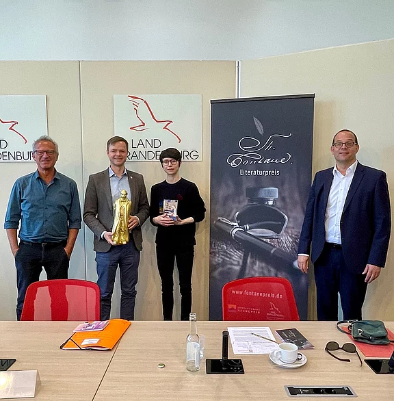Vier Personen stehen vor Bannern „Land Brandenburg“ und „Fontane Literaturpreis“. Eine Person hält eine goldene Figur, eine andere ein Buch; es wirkt wie eine Preisverleihung. Konferenzraum mit Tisch im Vordergrund.