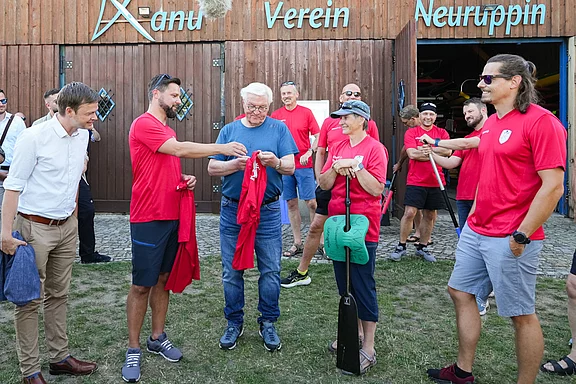 Eine Gruppe von Personen, darunter Männer und Frauen in roten T-Shirts, steht vor einem Holzgebäude. Sie interagieren freundlich miteinander, während einer von ihnen ein T-Shirt überreicht. Im Hintergrund sind weitere Teilnehmer sichtbar, die an der Veranstaltung teilnehmen.