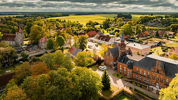 Luftaufnahme eines kleinen Dorfes mit verschiedenen Häusern und Bäumen. Im Vordergrund steht ein historisches Gebäude mit Türmchen, umgeben von einer grünen Landschaft und Feldern. Der Himmel ist bewölkt und die Farben sind herbstlich.