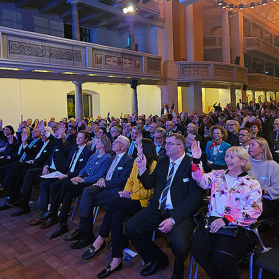 Eine große Menschenmenge sitzt in einem Saal und hebt ihre Hände, um abzustimmen oder Fragen zu stellen. Die Anwesenden tragen formelle Kleidung und scheinen an einer Veranstaltung teilzunehmen. Der Raum hat hohe Decken und eine festliche Atmosphäre.