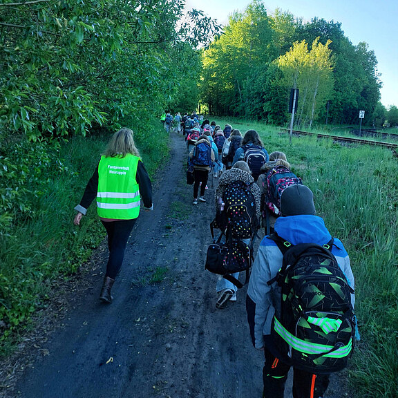 Eine Gruppe von Kindern und Jugendlichen geht auf einem Waldweg. Einige tragen Rucksäcke, während eine Person in einer grünen Warnweste die Gruppe anführt. Die Umgebung besteht aus Bäumen und Büschen, und im Hintergrund sind Gleise zu sehen.