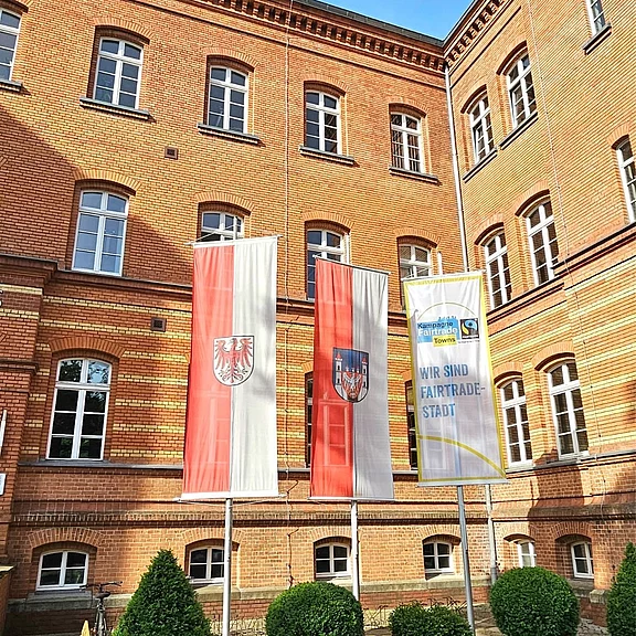 Im Hof eines historischen Backstein-Gebäudes stehen drei Fahnenmasten: zwei rot‑weiße Fahnen mit Wappen, daneben ein weiß-gelbes Banner mit Fairtrade-Logo und der Aufschrift „Wir sind Fairtrade-Stadt“.