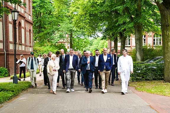 Eine Gruppe von Menschen geht auf einem gepflasterten Weg, umgeben von Bäumen und historischen Gebäuden. Die Teilnehmer tragen verschiedene Business-Outfits und scheinen an einem offiziellen Termin oder einer Besprechung teilzunehmen.