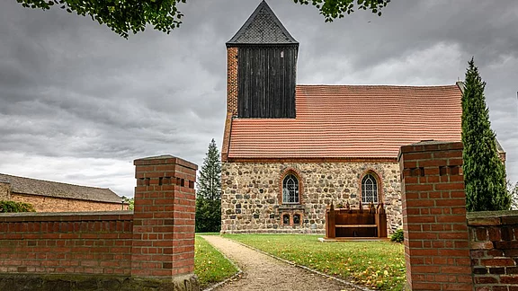 Eine Steinkirche mit einem roten Ziegeldach und einem schwarzen Holzschindelturm. Der Eingang wird von zwei Ziegelmauern flankiert, und der Weg zur Kirche ist von Bäumen gesäumt. Der Himmel ist bewölkt, was der Szene eine ruhige Atmosphäre verleiht.