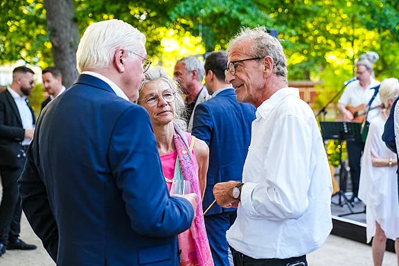 Drei Personen stehen in einem Gespräch in einem grünen, sonnigen Garten. Eine Frau mit grauen Haaren trägt eine rosa Schärpe. Zwei Männer, einer mit Brille und grauen Haaren, unterhalten sich mit ihr. Im Hintergrund sind unscharf weitere Gäste und eine kleine Bühne zu sehen.