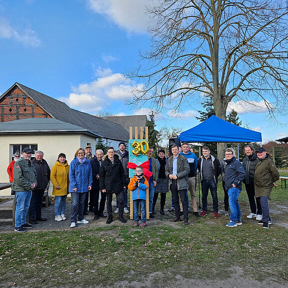 Eine Gruppe von etwa 20 Personen steht im Freien, umgeben von Bäumen und einem Gebäude. In der Mitte hält ein Kind eine Figur mit der Zahl 30. Im Hintergrund ist ein blauer Pavillon zu sehen. Die Stimmung wirkt festlich und gemeinschaftlich.