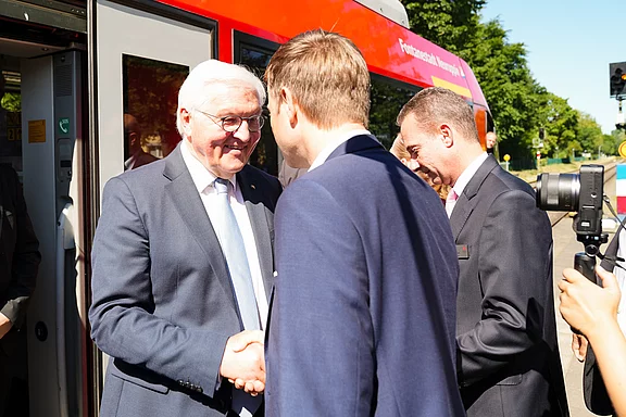 Der Bundespräsident lächelt und schüttelt die Hand eines Mannes vor einem roten Zug. Im Hintergrund sind weitere Personen sichtbar, die an einem Empfang teilnehmen. Die Szene vermittelt eine freundliche und offizielle Atmosphäre.