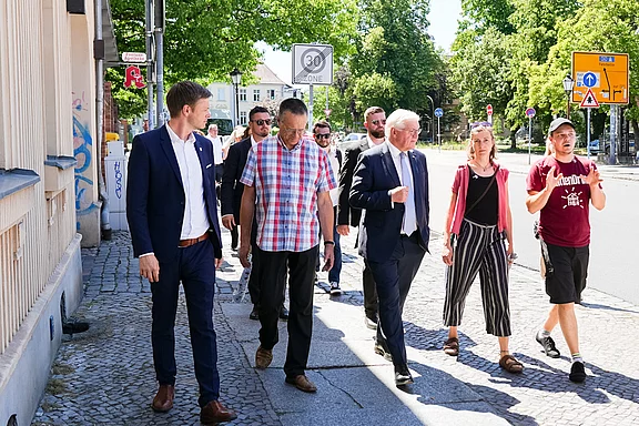 Eine Gruppe von Personen geht gemeinsam eine Straße entlang. Einige tragen formelle Kleidung, während andere leger gekleidet sind. Sie scheinen an einer Diskussion oder einem Gespräch beteiligt zu sein, während sie sich in einer städtischen Umgebung bewegen.