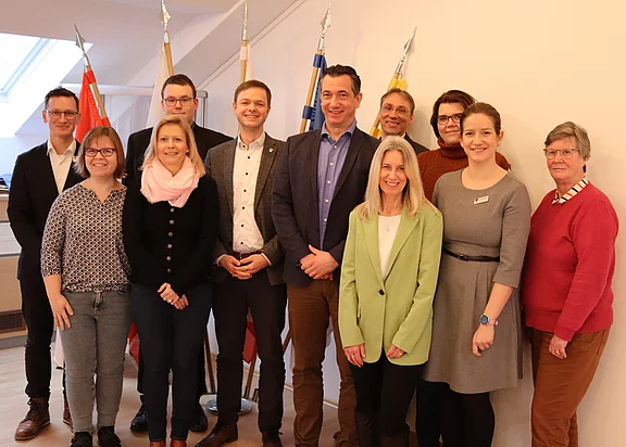 Eine Gruppe von elf Personen steht in einem hellen Raum, einige halten die Hände in den Hüften oder greifen in die Taschen. Im Hintergrund sind mehrere Fahnen zu sehen. Die Personen lächeln und wirken freundlich. Es scheint sich um ein Treffen oder eine Veranstaltung zu handeln.
