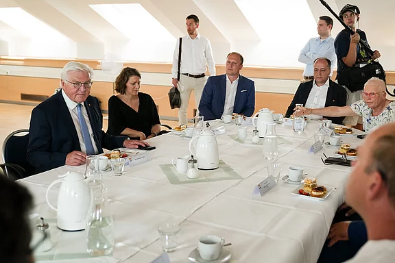 Eine Gruppe von Menschen sitzt an einem Tisch, umgeben von Teekannen, Gläsern und Snacks. Im Vordergrund spricht ein älterer Mann mit Brille, während andere Teilnehmer interessiert zuhören. Die Atmosphäre ist formal, wahrscheinlich handelt es sich um eine Diskussion oder ein Treffen.