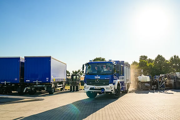 Ein blauer Einsatzwagen des Technischen Hilfswerks (THW) fährt auf einem Platz mit anderen Lastwagen und Container. Die Sonne scheint, und der Himmel ist klar. Die Umgebung wirkt industriell mit verschiedenen Fahrzeugen und Ausrüstungen im Hintergrund.