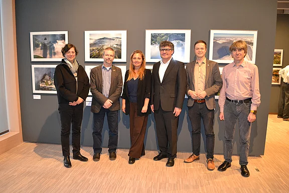 Sechs Erwachsene stehen nebeneinander in einer Galerie vor einer Wand mit Landschaftsfotografien. Sie posieren für ein Gruppenfoto, lächeln und tragen überwiegend formelle Kleidung. Ruhige Ausstellungsszene.