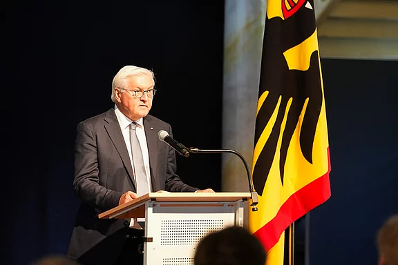 Ein Mann mit Brille in einem Anzug steht an einem Rednerpult und spricht. Hinter ihm befindet sich die deutsche Flagge. Der Fokus liegt auf dem Redner, der offenbar eine Ansprache hält.