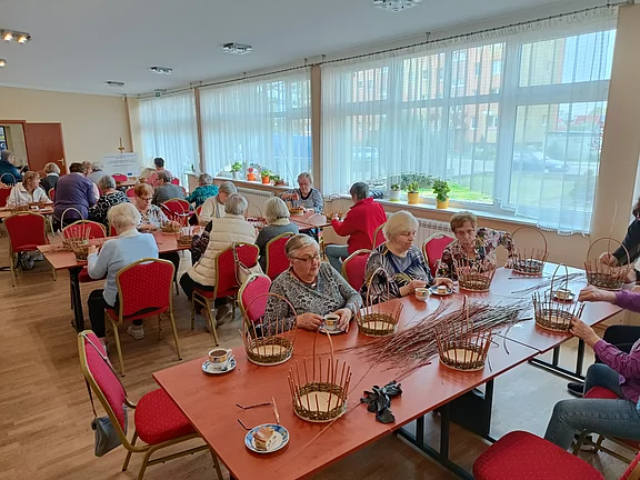 Mehrere ältere Menschen sitzen in einem hellen Gemeinschaftsraum an Tischen und nehmen an einem Bastel-Workshop teil. Sie flechten Weidenkörbe und trinken Kaffee. Freundliche, ruhige Atmosphäre.