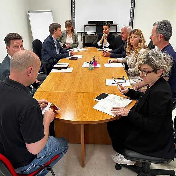 Gruppe von Menschen in einem Besprechungsraum sitzt um einen langen Holztisch. Sie diskutieren konzentriert, machen Notizen und tauschen Dokumente aus. Auf dem Tisch liegen Papiere, Stifte und Smartphones.