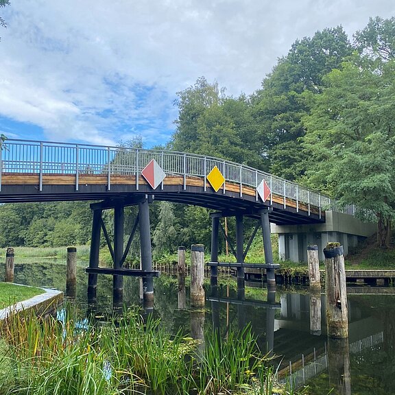 Fußgängerbrücke mit Metallgeländer über einem ruhigen Kanal, umgeben von dichtem Grün. Mehrere rautenförmige Schilder hängen an der Brücke. Schwarze Stützen und alte Holzpfähle ragen aus dem Wasser, das die Brücke spiegelt. Bewölkter Himmel.