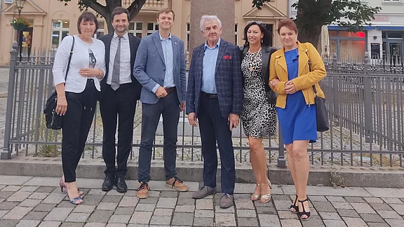 Sechs Erwachsene posieren lächelnd vor einer großen Statue auf einem städtischen Platz. Sie tragen überwiegend elegante Kleidung. Hinter ihnen stehen Bäume und ein umzäunter Sockel der Statue. Die Szene wirkt wie ein Erinnerungsfoto bei einem Besuch oder Termin.