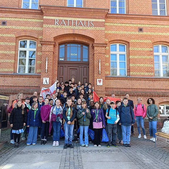 Große Gruppe Jugendlicher und Begleitpersonen posiert auf der Treppe vor einem Backstein-Rathaus. Über dem Eingang steht „RATHAUS“. Einige halten Fahnen. Es wirkt wie ein Klassen- oder Gruppenfoto bei einem Besuch der Stadtverwaltung.