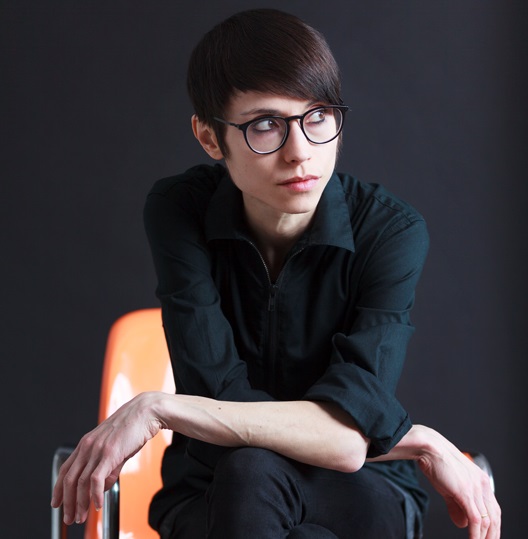 Ein junger Mann sitzt auf einem Stuhl, trägt ein schwarzes Hemd und eine Brille. Er hat kurze, glatte Haare und schaut nachdenklich zur Seite. Der Hintergrund ist dunkel, wodurch der Fokus auf seinem Gesicht und seiner Pose liegt.