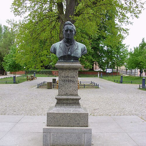 Bronzene Büste eines Mannes auf hohem Granitsockel, zentral auf einem gepflasterten Platz. Im Hintergrund Bäume und Bänke. Das Denkmal steht im Mittelpunkt der Szenerie.